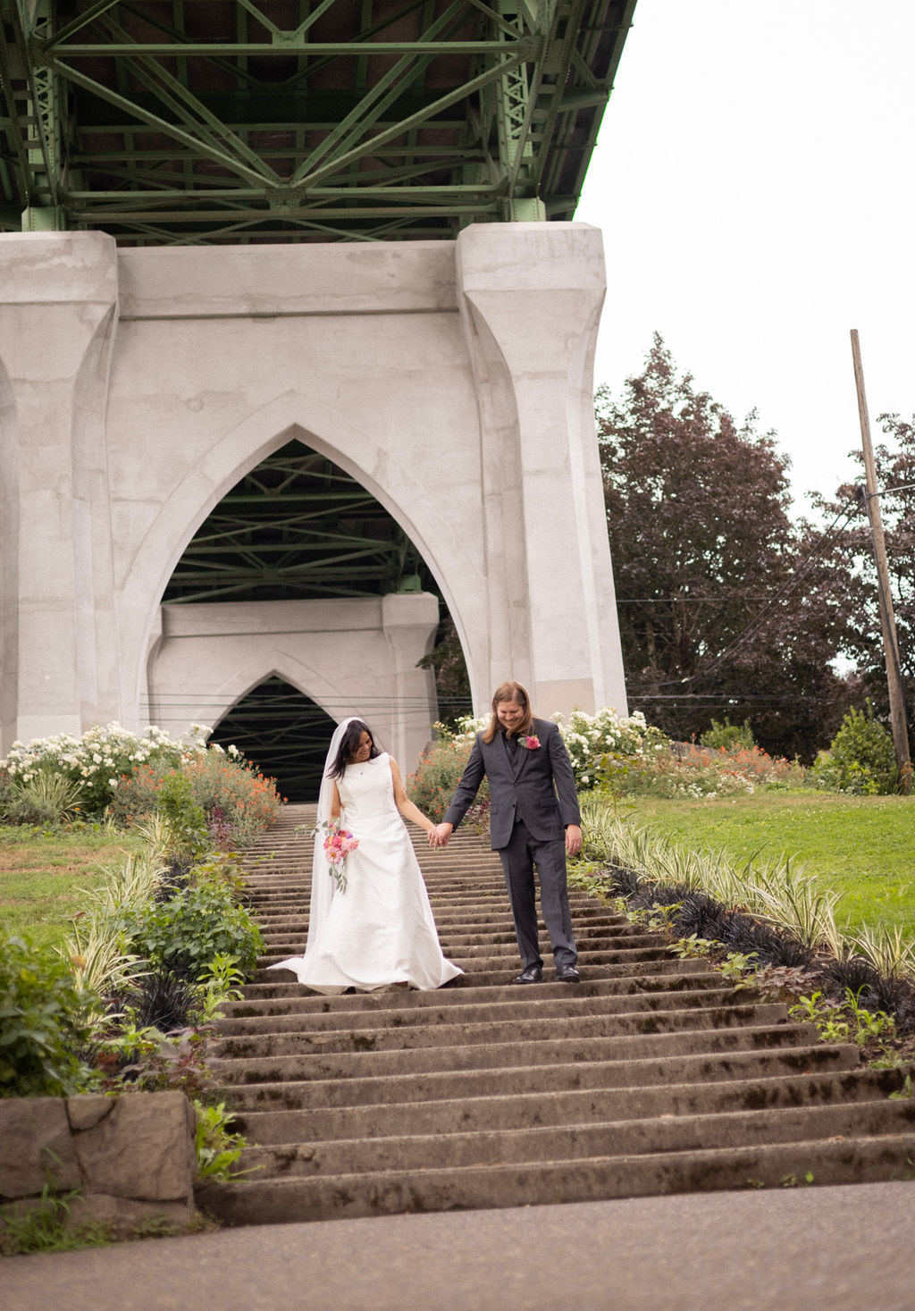 A couple walking down the Cathedral Park staircase under the gothic arch of the St. Johns Bridge after their ceremony