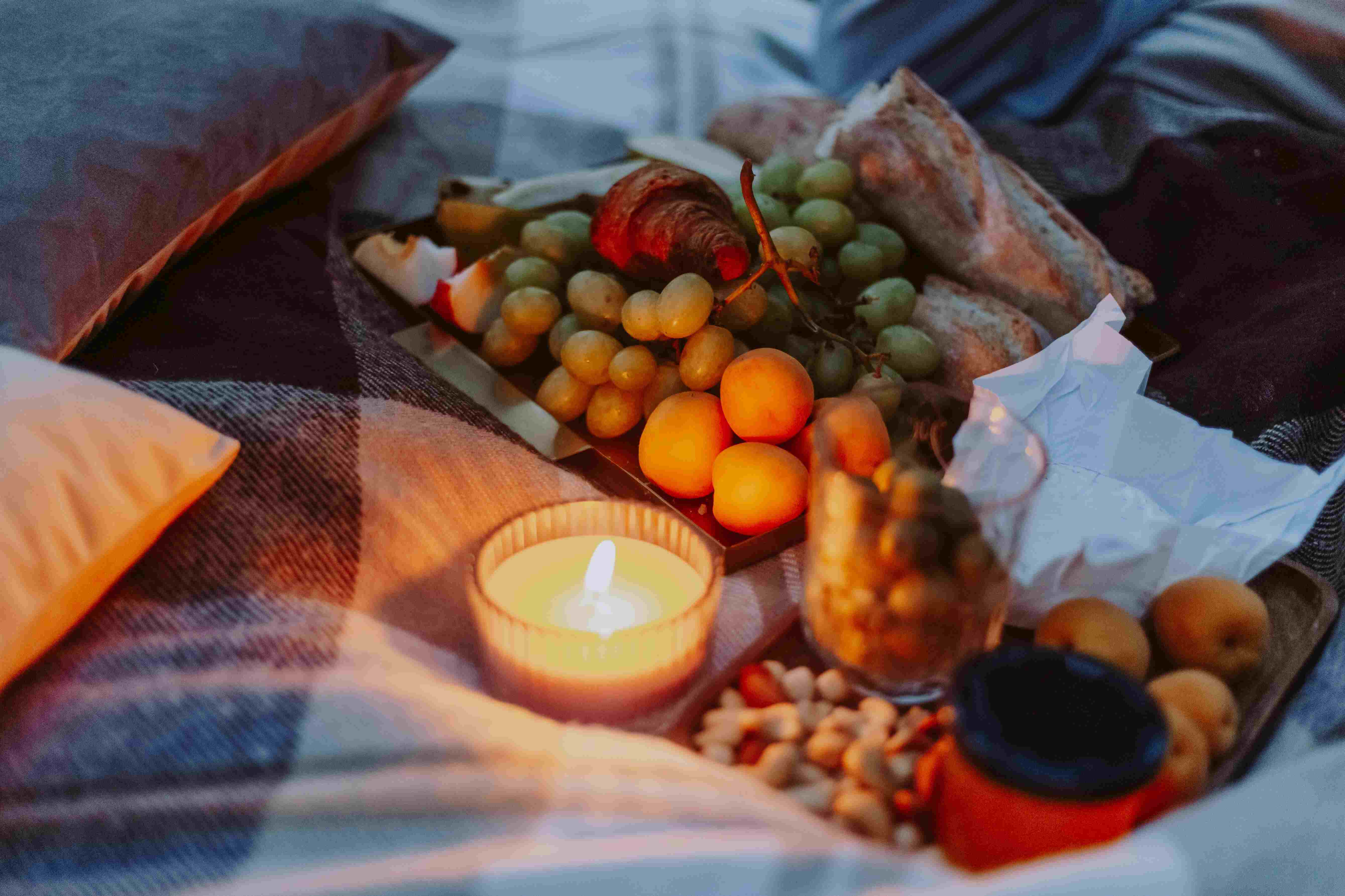 A candlelit picnic tray of fruit, bread and nuts on blankets and cushions at dusk