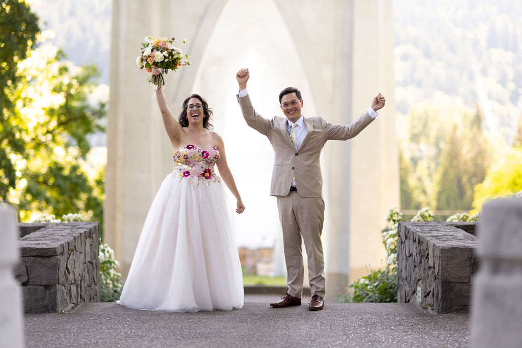A newly married couple cheering with raised arms beneath the arches of the St. Johns Bridge