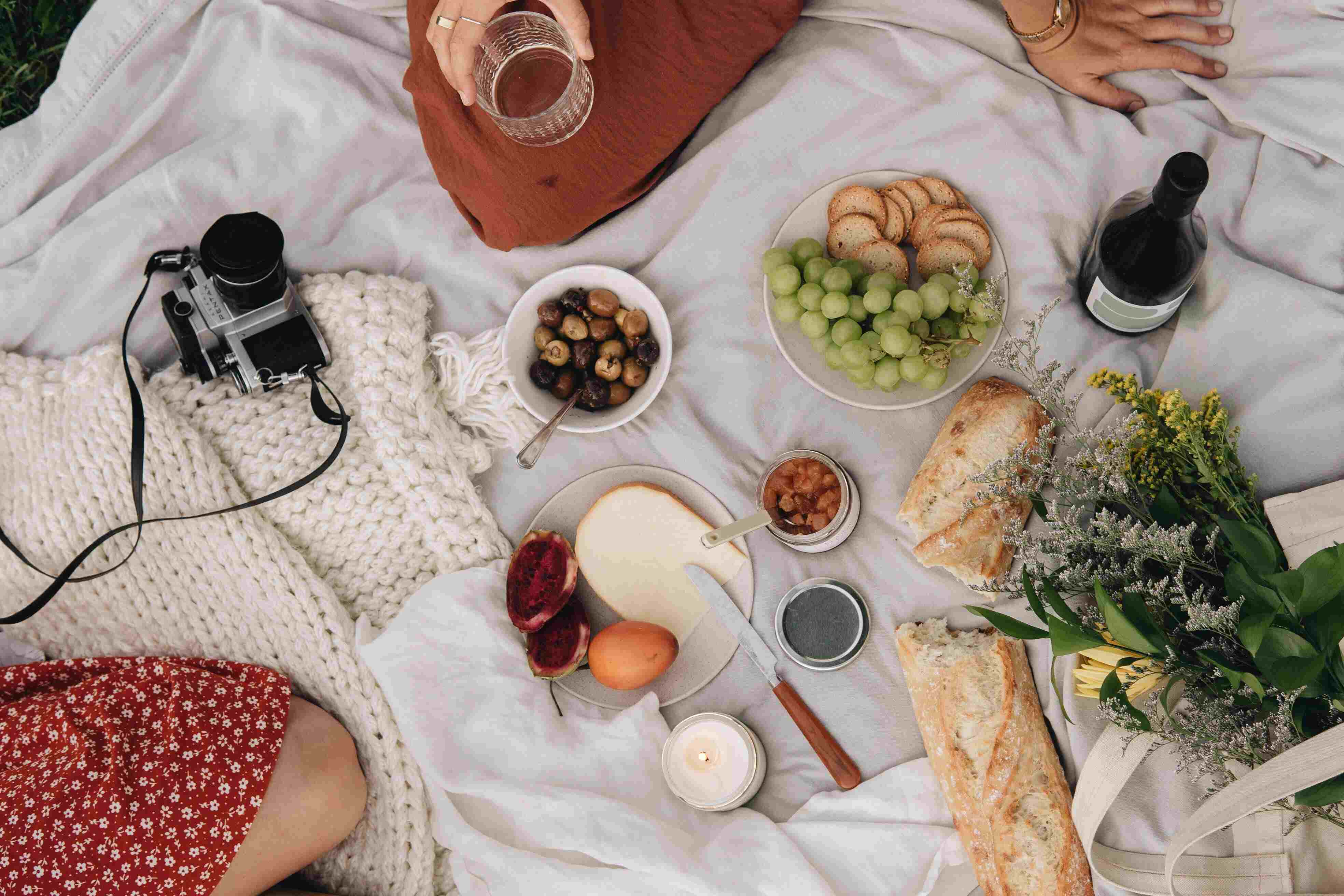 An overhead view of a blanket picnic with cheese, grapes, baguette, wine and wildflowers