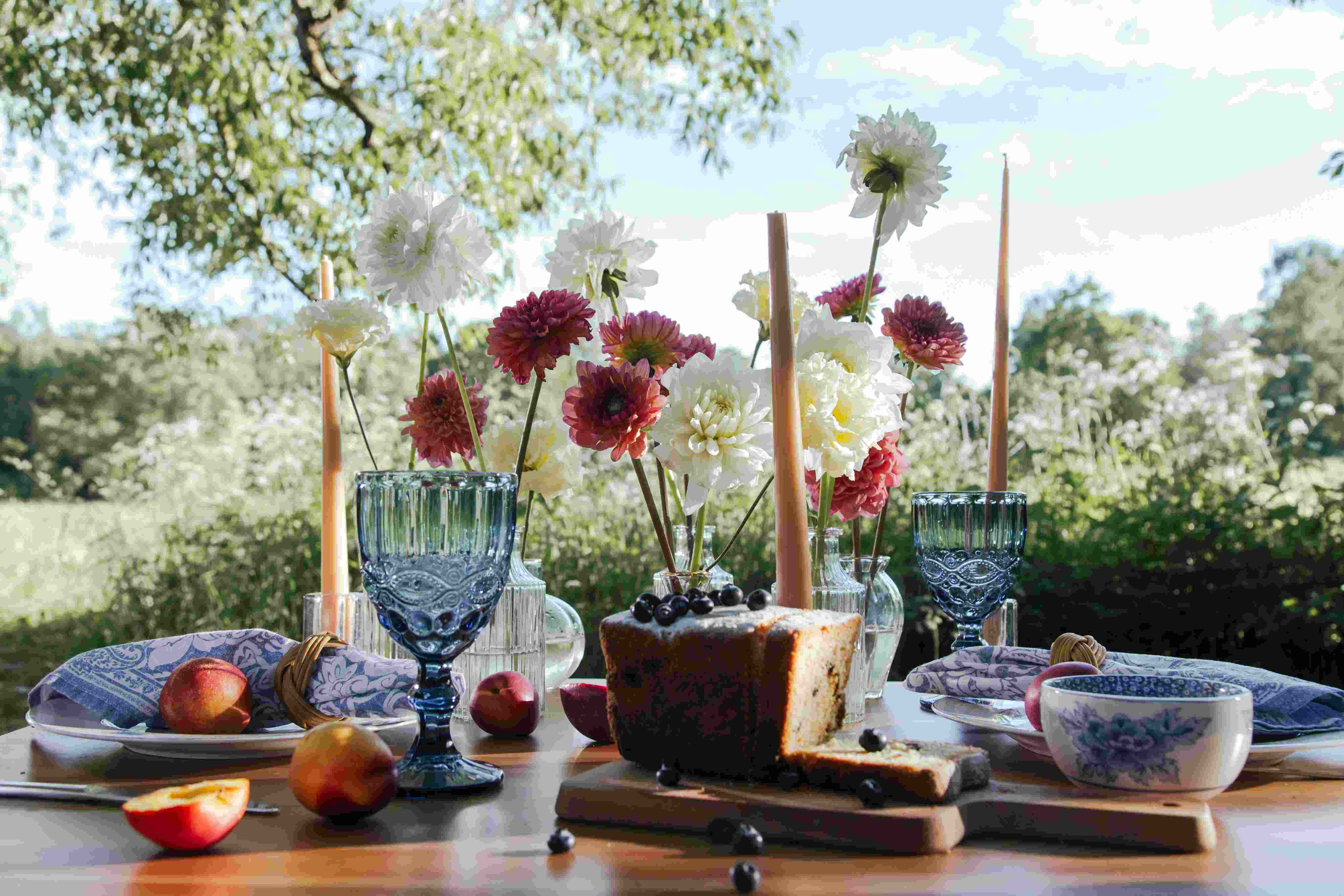A styled picnic table with dahlias, taper candles, stone fruit and a sliced loaf, set outdoors